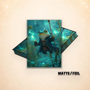 Op de afbeelding: Een fantasie-illustratie van een kikker-tovenaar in een blauwe jas, met een staf en het uitspreken van een spreuk met gloeiende groene energie. De afbeelding is gelabeld "MATTE/FOIL". De kikker bevindt zich in een waterige omgeving.
