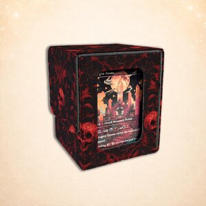 Op de afbeelding: Een vierkante deckbox met een zwart en rood patroon van schedels en bloemranken. De voorzijde heeft een rechthoekig uitgesneden venster waarin een ruilkaart te zien is met een maanverlichte landschapsillustratie en de tekst Savai Triome.