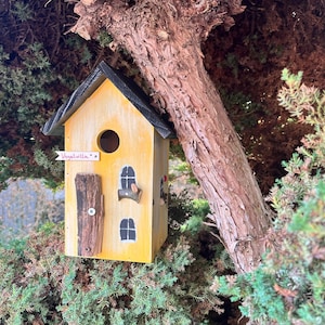 Könnte beinhalten: Ein gelbes Vogelhaus mit schwarzem Dach und Holztür befindet sich in einem Baum. Das Vogelhaus hat ein rundes Einflugloch und zwei kleine Fenster. Der Text "Vogelvilla" steht auf einem kleinen Banner. Das Vogelhaus ist von grünem Laub umgeben.