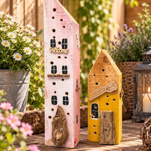Könnte beinhalten: Zwei hohe, schmale Insektenhotels aus Holz in Hausform. Das größere ist hellrosa gestrichen, hat schwarz aufgemalte Fenster und ein Holzschild mit der Aufschrift "Welcome". Das kleinere ist gelb und trägt ein Holzschild mit "Hallo Frühling".