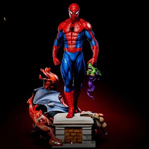 May include: A detailed figurine of Spider-Man in a classic red and blue suit, holding the Green Goblin's mask. The statue stands on a brick-like platform with other character elements, set against a dark background.