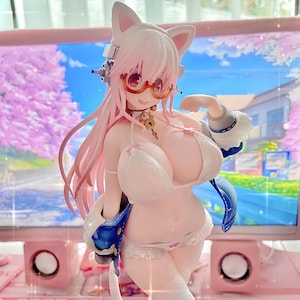 Peut inclure: Figurine de style anime rose et blanc représentant une femme avec des oreilles de chat, portant un bikini blanc, des lunettes et un casque. La figurine est posée devant un écran d'ordinateur et des haut-parleurs roses, avec un fond de fleurs de cerisier.