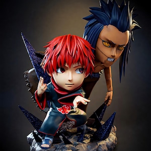 Op de afbeelding: Gedetailleerd beeldje met twee anime-personages. Eén personage heeft rood haar en draagt een zwarte outfit met rode accenten. De andere heeft blauw haar en een gebruinde teint. De figuren poseren op een rotsachtige basis met donkerblauwe, stekelige accenten.