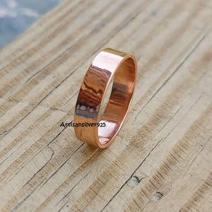 Può includere: Un anello di rame con una fascia piatta, appoggiato su una superficie di legno. L'anello ha una finitura lucida, che riflette la luce. L'anello è un design semplice e classico. Il testo "Artisansilver925" è visibile.
