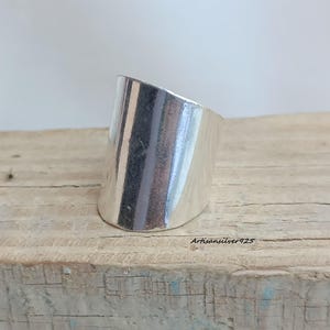 Pode incluir: Um anel largo prateado com um design inclinado. O anel é feito de um metal brilhante e tem uma superfície lisa. O anel é exibido em uma superfície de madeira. O texto "Artisansilver925" é visível na superfície de madeira.