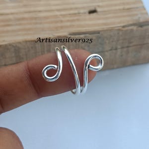 Könnte beinhalten: Ein silberfarbener Ring mit einem einzigartigen, offenen Design. Der Ring hat eine wirbelnde, kursive Form mit zwei abgerundeten Enden. Der Text "Artisansilver925" ist im Hintergrund sichtbar. Ein stilvolles Accessoire.