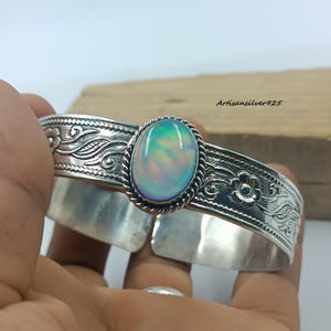 Pode incluir: Uma pulseira de punho de prata com uma pedra de opala oval. A pulseira apresenta intrincados padrões florais e de redemoinhos. A opala tem tons de azul, verde e rosa. O texto "Artisansilver925" é visível.