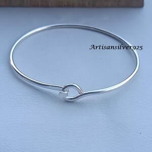 May include: A silver bangle bracelet with a hook and eye clasp. The bracelet is circular and has a smooth, polished finish. The text "Artisansilver925" is printed on the bracelet.