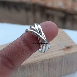 Könnte beinhalten: Ein silberner Ring mit einem gewebten Design. Der Ring hat mehrere miteinander verflochtene Bänder, die ein strukturiertes und kompliziertes Aussehen erzeugen. Der Ring wird auf einem Finger gezeigt, um seine Form und Details hervorzuheben. Der Ring ist aus Silber.