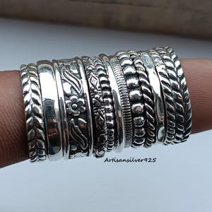 May include: A collection of silver rings with various designs, including floral, braided, and beaded patterns. The rings are stacked together, showcasing the different textures and styles. The rings are made of a shiny, metallic material.