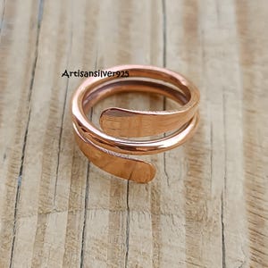 May include: A copper-toned ring featuring a spiral design. The ring coils around the finger twice, with the ends overlapping. The ring is presented on a wooden surface. The text "Artisansilver925" is visible.