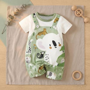 May include: A baby's romper set with a white t-shirt and green dungarees. The dungarees are patterned with a koala, giraffe, tiger, and other jungle animals. The romper has white buttons and a green trim.