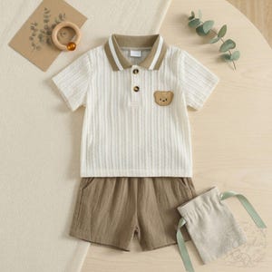 May include: A baby's two-piece outfit set. The white polo shirt has a brown collar and a bear patch. The shorts are brown, and a small drawstring bag is included. A wooden teething ring and a card with greenery are also in the image.