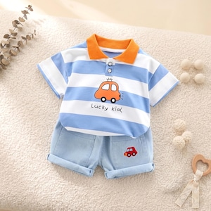 May include: A two-piece baby outfit featuring a blue and white striped polo shirt with an orange collar and a cartoon car graphic. The matching denim shorts have a car applique and rolled cuffs. The shirt says "Lucky kid".