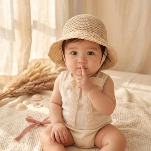 Könnte beinhalten: Ein Baby mit einem beigefarbenen Sonnenhut aus Stroh und einem ärmellosen Strampler mit einer Spitzenschleife. Das Baby sitzt auf einer cremefarbenen Decke und hält einen Finger an die Lippen. Der Hintergrund umfasst getrocknete Pflanzen und einen weichen Vorhang.