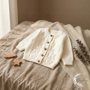 Puede incluir: Cárdigan de bebé color crema con diseño de punto trenzado y botones de madera. El suéter de manga larga se exhibe en una cama con juguetes de madera en forma de nube y estrella.