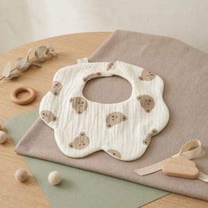 Könnte beinhalten: Ein cremefarbenes Baby-Lätzchen mit gewelltem Rand und einem Muster aus braunen Bärengesichtern. Das Lätzchen ist aus einem weichen, strukturierten Stoff gefertigt. Ein Beißring und ein Spielzeug aus Holz sind ebenfalls zu sehen.