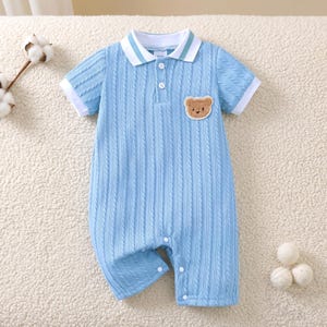 May include: Pale blue baby romper with a cable knit design. It has a white and light blue striped collar, short sleeves with white cuffs, and a small bear patch. The romper fastens with buttons.