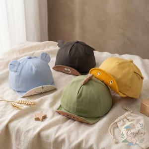 Op de afbeelding: Vier kleurrijke baby baseballpetten. Eén is lichtblauw met berenoren, een andere is donkergrijs met vleermuisvleugels, een derde is geel met een lachend gezicht en de laatste is groen met een dinosaurusvin. Elke pet heeft een contrasterende klep.