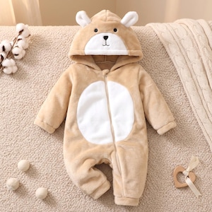 Peut inclure: Une combinaison une pièce à manches longues en peluche beige, dotée d'une capuche avec des oreilles d'ours et un visage d'ours. Le vêtement présente un panneau circulaire blanc sur la poitrine et une fermeture éclair centrale, en matière polaire douce.