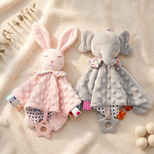 Könnte beinhalten: Zwei Baby-Schmusetücher. Eines ist rosa, in Form eines Hasen mit langen Ohren, und das andere ist grau, in Form eines Elefanten. Beide haben gemusterte Kragen, strukturierte Körper und sternförmige Beißringe.