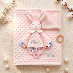 Può includere: Una coperta rosa per bambini con una testa e un corpo di coniglio di peluche. Il coniglio ha lunghe orecchie, occhi chiusi e un colletto a pois. La coperta ha una superficie strutturata e il nome "Olivia" è ricamato sopra.