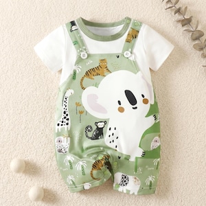 Op de afbeelding: Een babyromper met korte mouwen, bestaande uit een wit t-shirt en een aangehechte salopette in saliegroen. De groene stof is versierd met een grote witte koala-afbeelding, kleinere illustraties van een tijger, giraf, aap en palmbomen.