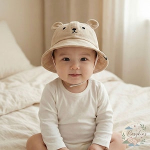 Könnte beinhalten: Beiger Baby-Eimerhut mit Bärenohren und einem aufgestickten Bärengesicht. Der Hut hat einen Kinnriemen und wird von einem Baby getragen, das ein weißes Langarmshirt trägt. Das Baby sitzt auf einem Bett.