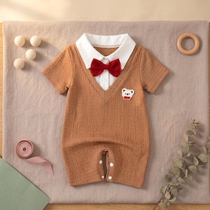 Puede incluir: Mono de bebé marrón con cuello blanco, pajarita roja y aplique de oso. El mono de manga corta tiene cuello en V y cierre de botones. La tela es texturizada. Ideal para bebés.