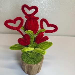 May include: A handcrafted, decorative floral arrangement featuring a red rose and three red heart accents. The flower and hearts are made of a fuzzy material, with green leaves and a green base, all set in a brown pot.