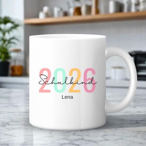 Könnte beinhalten: Eine weiße Keramiktasse mit dem farbigen Text 2026 in Pink, Mintgrün und Gelb. Darüber steht in schwarzer Schreibschrift das Wort Schulkind, wobei der Name Lena in einer einfachen schwarzen serifenlosen Schrift direkt unter dem Jahr gedruckt ist.