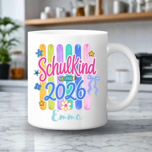 Könnte beinhalten: Ein weißer Keramikbecher mit einer bunten Grafik aus vertikalen Streifen in Orange, Gelb, Lila, Türkis, Blau und Pink. Der Text lautet Schulkind 2026 und Emma, ergänzt durch Illustrationen eines Bleistifts, Sternen, Blumen und einer blauen Schleife.