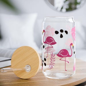 Könnte beinhalten: Ein durchsichtiges Glas in Dosenform mit einem Muster aus rosa Flamingos, tropischen Blättern und schwarzen Punkten. Das Set enthält einen runden Bambusdeckel mit einem mittigen Loch sowie einen geraden Trinkhalm aus klarem Glas.