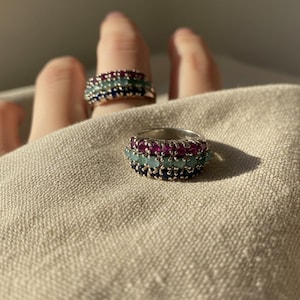 Può includere: Due anelli d'argento con pietre preziose colorate. Un anello è su un dito e l'altro è in primo piano. Gli anelli hanno tre file di pietre preziose nei toni del rosso, blu e verde. Gli anelli sono su uno sfondo di tessuto neutro.