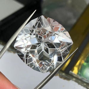 May include: A clear, colorless gemstone with a complex, multi-faceted geometric cut. The stone features a symmetrical, star-like pattern of triangular and trapezoidal facets that reflect light, held by a pair of metal tweezers for display.
