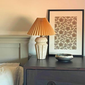 May include: A table lamp with a pleated light orange shade and a textured, speckled beige ceramic base with a geometric, multi-faceted design. Next to the lamp is a small, shallow, dark-toned ceramic bowl with a smooth, rounded rim.
