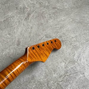 K&ouml;nnte beinhalten: Nahaufnahme eines Gitarrenhalses mit geflammter Ahorn-Oberfl&auml;che. Die Kopfplatte hat sechs L&ouml;cher f&uuml;r die Stimmwirbel. Die Holzmaserung hat eine warme, bernsteinfarbene Farbe mit gl&auml;nzendem Finish.