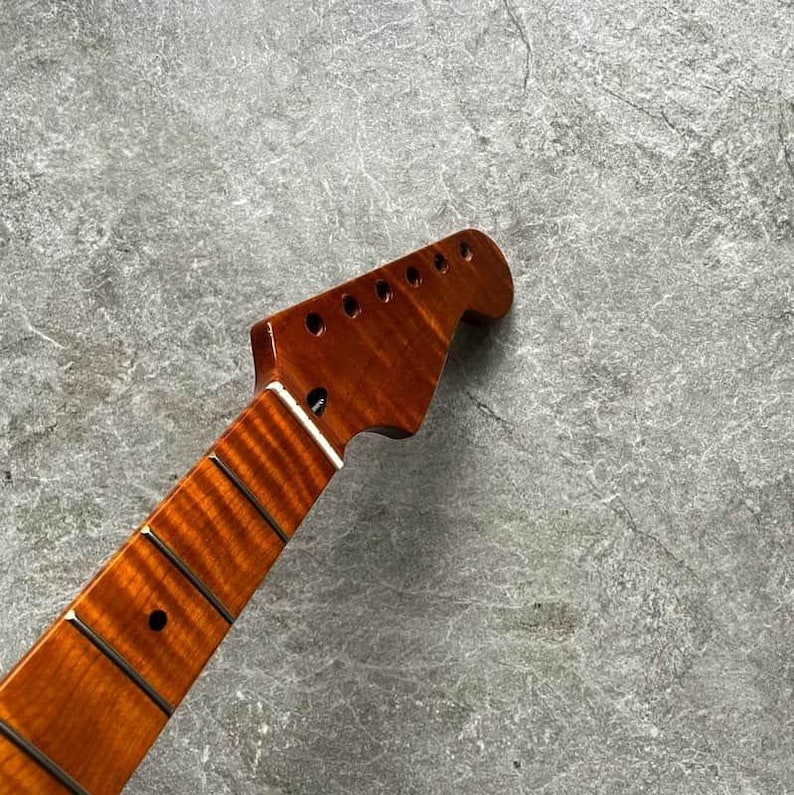 May include: A close-up of a guitar neck with a rich, reddish-brown finish. The headstock features six tuning peg holes. The fretboard has dark frets and inlaid position markers. The wood grain is visible, adding to the instrument's aesthetic.