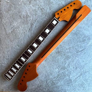 Puede incluir: Dos mástiles de guitarra sin terminar. Uno presenta un diapasón oscuro con incrustaciones blancas y una pala con agujeros para clavijas. El otro es de madera más clara con una franja oscura en la parte posterior y una pala con agujeros para clavijas.