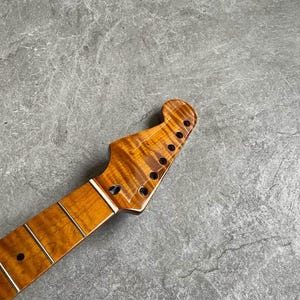 K&ouml;nnte beinhalten: Gitarrenhals mit geflammter Ahorn-Oberfl&auml;che. Die Kopfplatte hat sechs L&ouml;cher f&uuml;r die Stimmwirbel. Das Griffbrett hat eingelegte B&uuml;nde und einen wei&szlig;en Sattel. Die Holzmaserung hat eine warme, bernsteinfarbene Farbe.