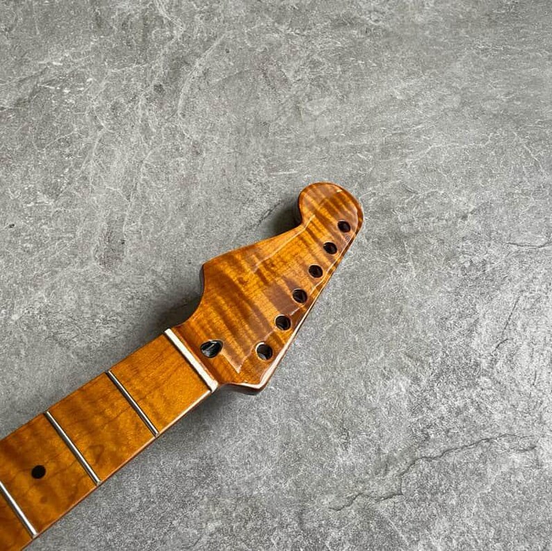 K&ouml;nnte beinhalten: Gitarrenhals mit geflammter Ahorn-Oberfl&auml;che. Die Kopfplatte hat sechs L&ouml;cher f&uuml;r die Stimmwirbel. Das Griffbrett hat eingelegte B&uuml;nde und einen wei&szlig;en Sattel. Die Holzmaserung hat eine warme, bernsteinfarbene Farbe.