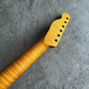 May include: A close-up of a guitar neck, crafted from light-colored wood with a prominent grain pattern. The headstock features six tuning peg holes. The wood has a warm, amber hue, with darker streaks adding depth and detail to the design.