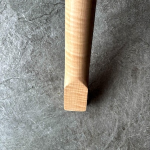 May include: Close-up of a light-colored wooden object with a rectangular end. The wood grain is visible, showing a natural pattern. The object appears to be a handle or part of a larger item.