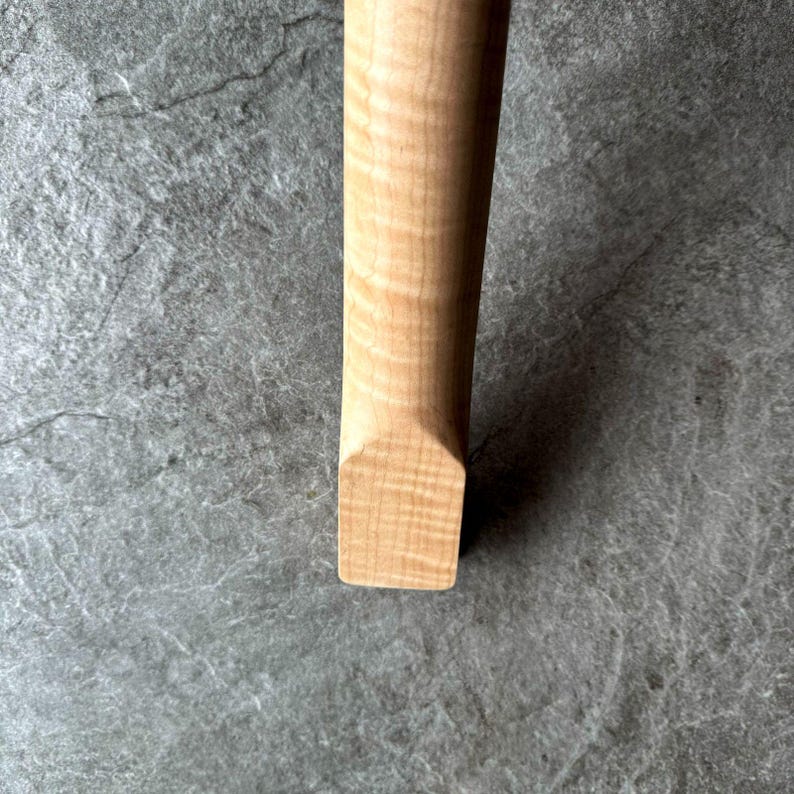 May include: Close-up of a light-colored wooden object with a rectangular end. The wood grain is visible, showing a natural pattern. The object appears to be a handle or part of a larger item.
