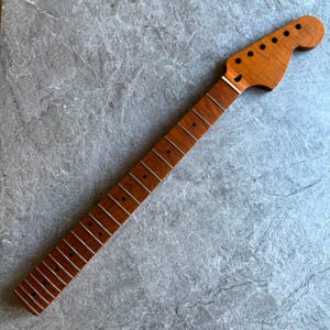 May include: A guitar neck made of light brown wood, featuring a headstock with tuning peg holes and a fretboard with inlaid frets. The neck has a natural wood grain pattern and is designed for a stringed instrument.