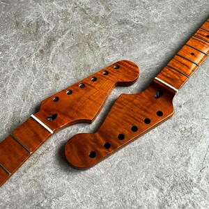 May include: Two unfinished guitar necks with a flamed maple grain pattern. The necks are a warm amber color, with pre-drilled holes for tuning pegs and frets. The headstocks have a classic shape.