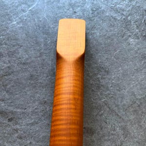 May include: Close-up of a wooden guitar neck with a light brown finish. The wood grain is visible, with a darker, wavy pattern running along the length of the neck. The headstock end is a lighter shade of brown.