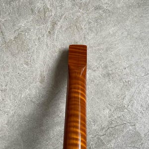 K&ouml;nnte beinhalten: Nahaufnahme eines h&ouml;lzernen Gitarrenhalses mit gl&auml;nzendem, bernsteinfarbenem Finish. Die Holzmaserung ist sichtbar und erzeugt ein gestreiftes Muster. Der Hals hat ein rechteckiges Ende und verj&uuml;ngt sich. Der Hintergrund ist ein meliertes Grau.