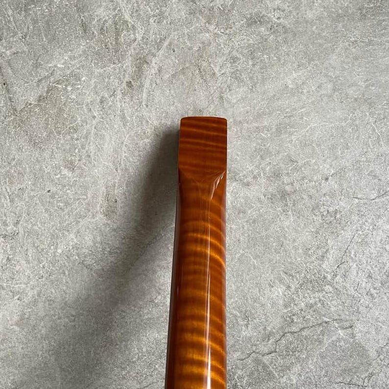 K&ouml;nnte beinhalten: Nahaufnahme eines h&ouml;lzernen Gitarrenhalses mit gl&auml;nzendem, bernsteinfarbenem Finish. Die Holzmaserung ist sichtbar und erzeugt ein gestreiftes Muster. Der Hals hat ein rechteckiges Ende und verj&uuml;ngt sich. Der Hintergrund ist ein meliertes Grau.