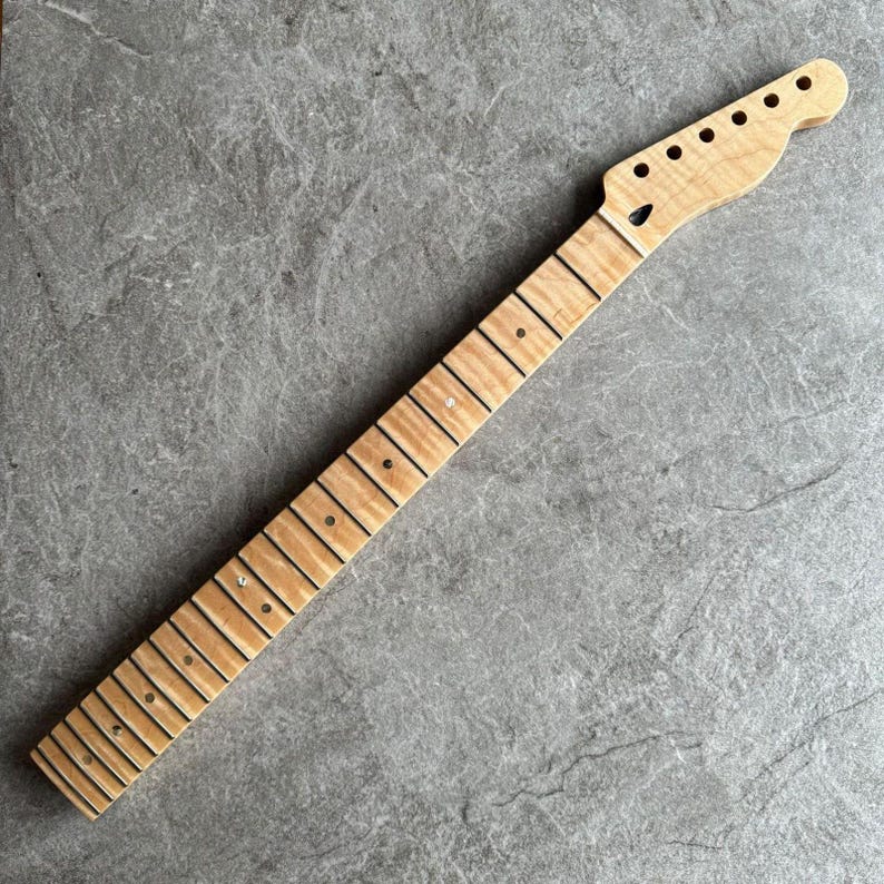 May include: A light-colored, unfinished guitar neck with a headstock featuring six tuning peg holes. The fretboard has dark lines and dot inlays. The wood grain is visible throughout the neck.